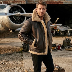 May include: A brown leather aviator jacket with a tan shearling collar and lining. The jacket has a classic design with a zip closure and pockets. The person is wearing black jeans. The background includes vintage airplanes and a hangar.