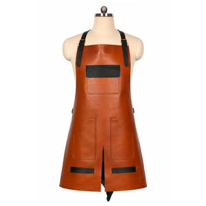 Könnte beinhalten: Eine hellbraune Schürze mit schwarzen Riemen und verstellbaren Schnallen, einer Brusttasche mit schwarzer Klappe, zwei großen unteren Taschen und zwei horizontalen schwarzen Zierstreifen am Saum, präsentiert auf einem Schneiderbüsten-Torso.