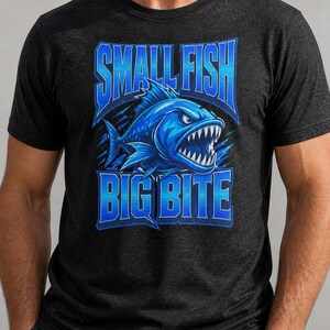 以下が含まれることがあります： 鋭い歯を持つ魚の青いイラストが描かれた濃い灰色のTシャツ。「SMALL FISH BIG BITE」の文字が青色の様式化されたフォントで表示され、大胆なグラフィックデザインを作成しています。