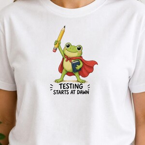 Puede incluir: Camiseta blanca con un dibujo de una rana superhéroe sosteniendo un lápiz y un libro. La rana lleva una capa roja y una bufanda roja. El texto "TESTING STARTS AT DAWN" está impreso debajo en negro.