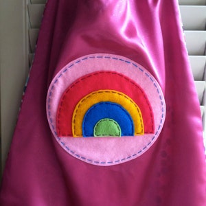 May include: A pink satin cape with a rainbow applique. The rainbow is made of felt and has red, orange, yellow, blue, green, and pink colors.