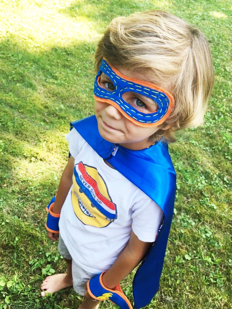 May include: A young child dressed as a superhero wearing a blue cape, a blue and orange mask, and blue and orange arm bands. The child is wearing a white t-shirt with the words "Never Give Up" printed on it.