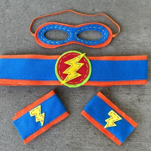 May include: A superhero costume set for kids, featuring a blue and orange felt mask, a blue and orange felt armband, and a blue and orange felt headband with a yellow lightning bolt.