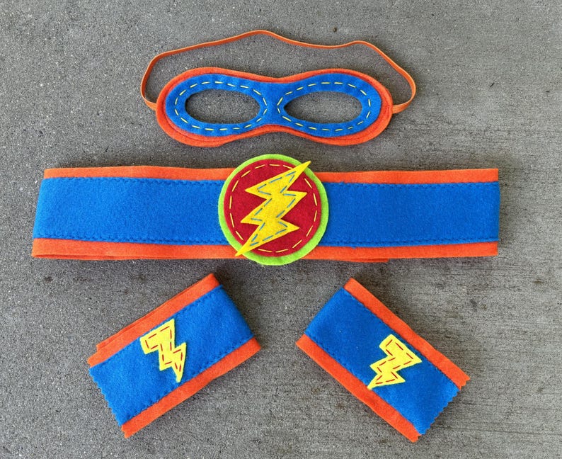May include: A superhero costume set for kids, featuring a blue and orange felt mask, a blue and orange felt armband, and a blue and orange felt headband with a yellow lightning bolt.