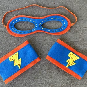 May include: A blue and orange felt superhero costume set with a mask and two arm bands. The arm bands have a yellow lightning bolt design.