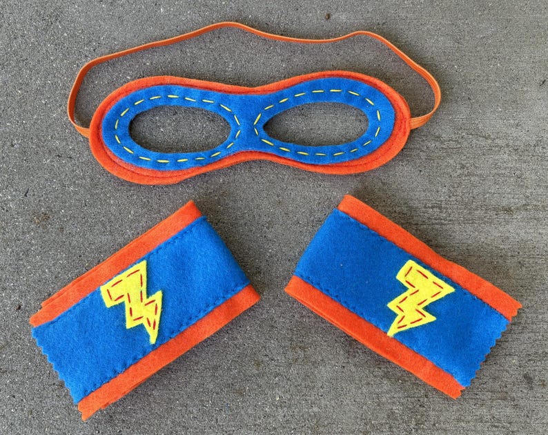 May include: A blue and orange felt superhero costume set with a mask and two arm bands. The arm bands have a yellow lightning bolt design.