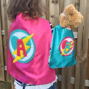 May include: Two superhero capes, one pink and one teal, with felt appliques. The pink cape has a yellow lightning bolt and the letter "A" in red. The teal cape has a yellow lightning bolt and the letter "B" in red.