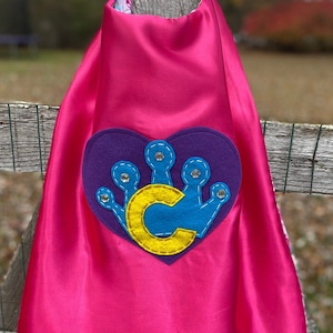 May include: Pink satin cape with a purple felt heart and a blue crown with a yellow letter C in the center. The crown has rhinestones on the points.