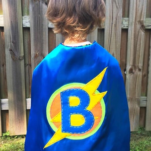 May include: A blue superhero cape with a yellow lightning bolt and a yellow letter "B" in the centre. The cape is decorated with orange, green, and blue stitching.