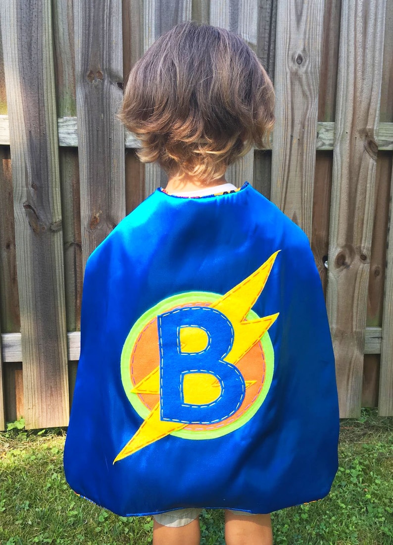 May include: A blue superhero cape with a yellow lightning bolt and a yellow letter "B" in the centre. The cape is decorated with orange, green, and blue stitching.