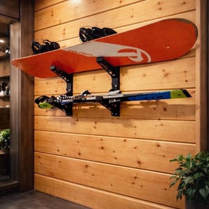 Puede incluir: Un snowboard y esquís montados en una pared de madera con soportes metálicos negros. El snowboard es naranja con detalles blancos y los esquís son azules y negros. La pared está hecha de tablones de madera horizontales.
