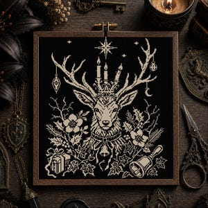 May include: A square cross-stitch embroidery in a dark wooden frame featuring a cream-colored deer head with large antlers adorned with three lit candles, hanging ornaments, a crescent moon, flowers, holly leaves, a gift box, and a hand bell.