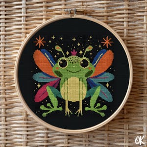 May include: A cross-stitch embroidery hoop featuring a whimsical frog with colorful wings, a crown, and star accents against a black background. The frog is green, yellow, and orange, with blue and pink wings. The hoop is set in a wooden frame.