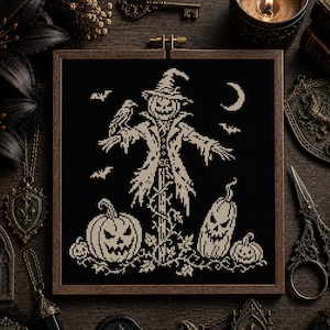 May include: A square wooden embroidery hoop containing a black fabric piece with a white cross-stitch design. The pattern features a central scarecrow with a pumpkin head, a crow on its shoulder, three jack-o'-lanterns, bats, and a crescent moon.