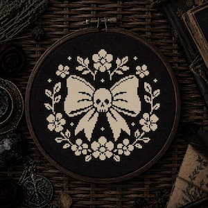 Puede incluir: Un bastidor de bordado circular de madera que contiene tela negra con un diseño de punto de cruz. El patrón presenta un lazo central de color crema con una calavera en el centro, rodeado por una corona simétrica de flores y hojas crema.
