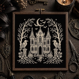 May include: A square embroidery hoop containing a cross-stitch pattern on black fabric. The design features a beige gothic mansion, two ravens perched on stone pillars, bare trees, pumpkins, and a crescent moon with bats in the night sky.