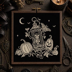 May include: A square wooden embroidery hoop containing a black fabric piece with a cream-coloured cross-stitch design. The pattern features a central lantern, a moth, pumpkins, a jack-o'-lantern, vines, a crescent moon, and small stars.