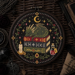 May include: A circular embroidery hoop containing a cross-stitch pattern on black fabric. The design features a green dragon sleeping on a stack of three books, surrounded by a candle, a potion bottle, floral vines, and a yellow crescent moon.