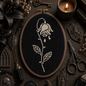 May include: An oval wooden embroidery hoop containing a cross-stitch pattern of a single drooping rose. The design is rendered in off-white thread against a solid black fabric background, featuring a stem with two leaves and a wilted flower head.