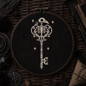 May include: A circular wooden embroidery hoop containing black fabric with a cross-stitch design of a skeleton key. The key features a bird skull at the top, a ribcage in the center, and four small star symbols surrounding the central key structure.