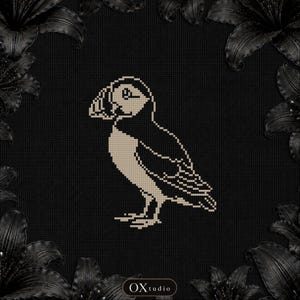May include: A cross-stitch pattern featuring a beige and black puffin bird centered on a black grid fabric. The design is framed by dark, glossy lily flowers in the corners. The text OXtudio is printed in a small white oval at the bottom center.
