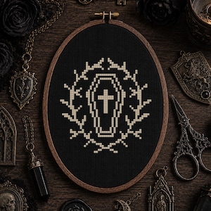 May include: An oval wooden embroidery hoop containing black fabric with a cross-stitched design in off-white thread. The design features a central coffin with a cross inside, surrounded by a symmetrical wreath of thorny branches.