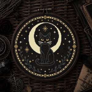 Peut inclure: Broderie au point de croix d'un chat noir devant un croissant de lune crème, entouré d'étoiles et de motifs floraux. Le motif est encadré dans un cercle en bois avec une bordure dorée en forme de chaîne.