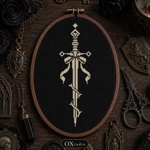 May include: An oval wooden embroidery hoop containing a black fabric piece with a cross-stitched design of a cream-colored sword adorned with a ribbon bow and wrapped in vines. The text OXtudio is printed in white at the bottom of the frame.