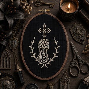 May include: An oval wooden embroidery hoop containing black fabric with a white cross-stitch design of a hand holding a cross, surrounded by a wreath. The design is centered within the hoop, which features a metal tightening screw at the top.