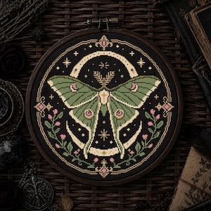 May include: A round, framed cross-stitch featuring a green moth with intricate details against a black background. The design includes a crescent moon, stars, and floral accents in pink and gold. The wooden frame adds a rustic touch to the artwork.