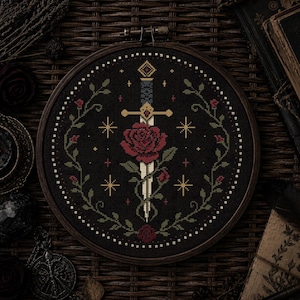 May include: Cross-stitch embroidery in a wooden hoop. The design features a sword piercing a red rose, surrounded by a floral wreath and stars, all on a black background. The embroidery is framed by a dark wooden hoop.