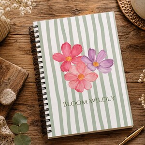 May include: A spiral-bound notebook featuring a light green and white vertical striped cover with a watercolor illustration of three flowers in shades of pink and purple, with the text BLOOM WILDLY printed in dark green lettering at the bottom.