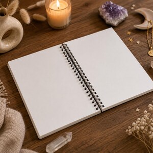 May include: An open spiral-bound notebook with blank white pages featuring a black metal coil spine. The notebook is positioned on a dark wooden surface, surrounded by a lit candle, a ceramic vase, a purple crystal, and a gold-toned necklace.