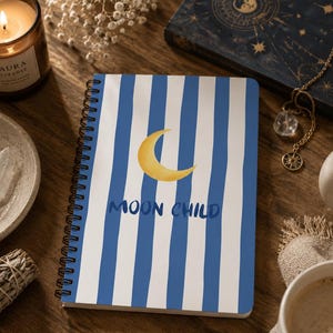 May include: A spiral-bound notebook featuring a white cover with vertical blue stripes, a yellow crescent moon illustration, and the text "MOON CHILD" in blue. The notebook is positioned on a wooden surface among various decorative items.