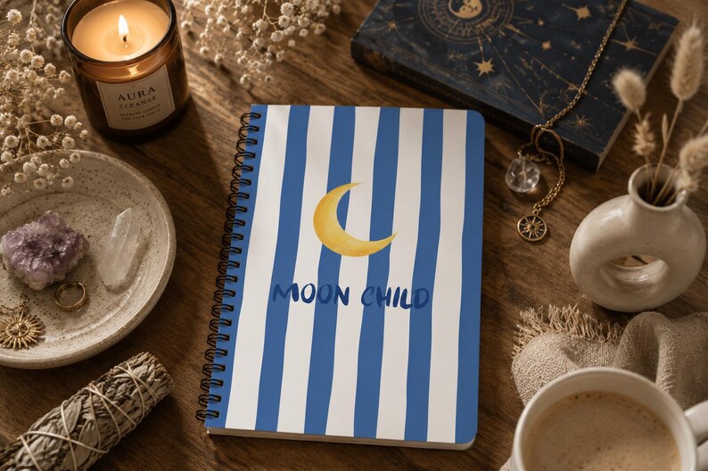 May include: A spiral-bound notebook featuring a white cover with vertical blue stripes, a yellow crescent moon illustration, and the text "MOON CHILD" in blue. The notebook is positioned on a wooden surface among various decorative items.