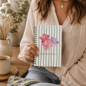 May include: A spiral-bound notebook featuring a light green and white vertical striped pattern. The cover displays a watercolor illustration of pink and purple flowers with the text BLOOM WILDLY printed in a dark green serif font below the flowers.