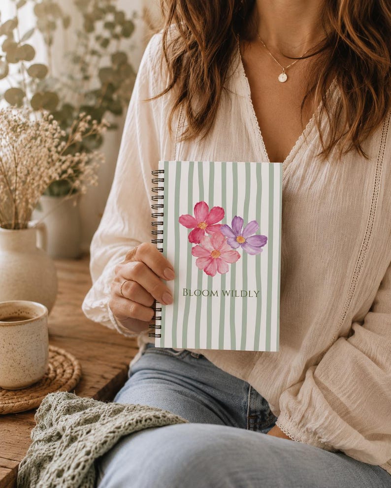 May include: A spiral-bound notebook featuring a light green and white vertical striped pattern. The cover displays a watercolor illustration of pink and purple flowers with the text BLOOM WILDLY printed in a dark green serif font below the flowers.
