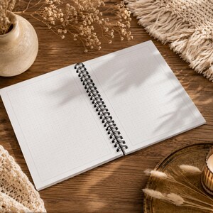 May include: An open spiral-bound notebook with white pages featuring a light gray dot grid pattern. The notebook has a black metal wire binding along the center fold, allowing the pages to lay flat for writing or drawing on the dotted surface.