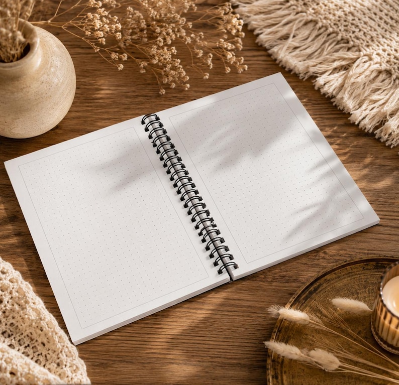 May include: An open spiral-bound notebook with white pages featuring a light gray dot grid pattern. The notebook has a black metal wire binding along the center fold, allowing the pages to lay flat for writing or drawing on the dotted surface.