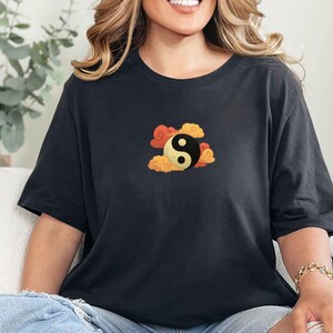 Może przedstawiać: Czarny t-shirt z symbolem Yin i Yang otoczonym pomarańczowymi i ż&oacute;łtymi chmurami. Grafika jest wyśrodkowana na klatce piersiowej. Koszulka jest w ciemnoszarym kolorze i ma kr&oacute;tkie rękawy.