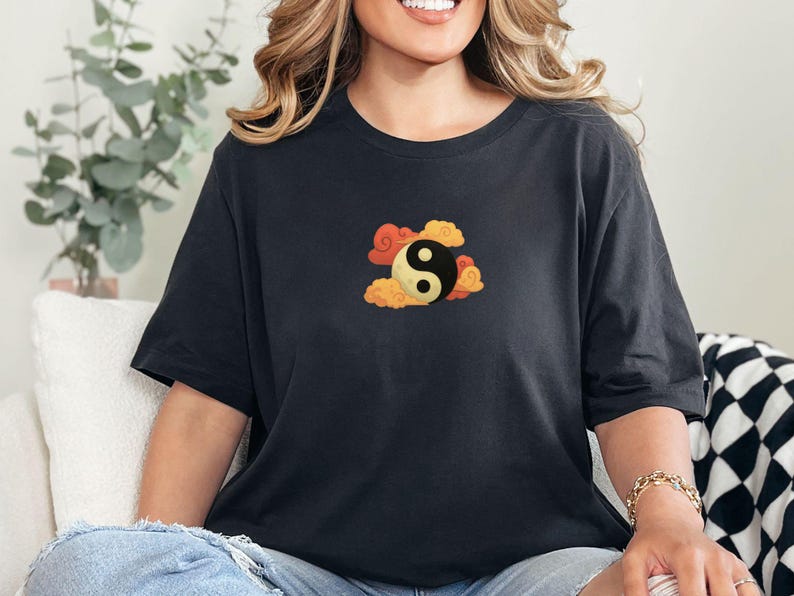 Może przedstawiać: Czarny t-shirt z symbolem Yin i Yang otoczonym pomarańczowymi i ż&oacute;łtymi chmurami. Grafika jest wyśrodkowana na klatce piersiowej. Koszulka jest w ciemnoszarym kolorze i ma kr&oacute;tkie rękawy.
