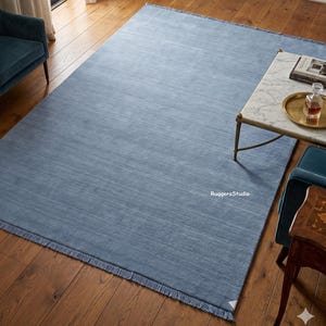 Pode incluir: Um tapete retangular de cor azul-claro sólida com uma textura horizontal subtil e franjas curtas ao longo das duas extremidades mais curtas. O tapete apresenta o texto RuqgersStudio impresso a branco na superfície do tecido.