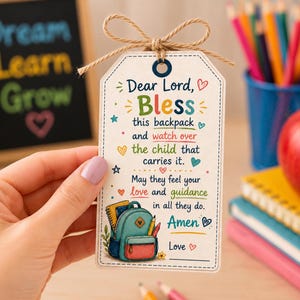 May include: A rectangular white gift tag with a jute twine bow at the top. The tag features colorful text that reads: Dear Lord, Bless this backpack and watch over the child that carries it. May they feel your love and guidance in all they do. Amen.