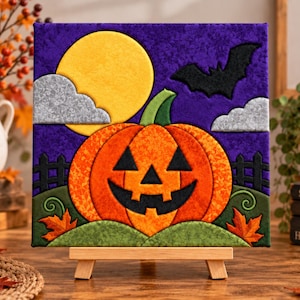 May include: A square fabric wall art piece featuring a central orange jack-o'-lantern with a black face, set against a purple background. The scene includes a yellow full moon, grey clouds, a black bat, a black fence, and orange autumn leaves.