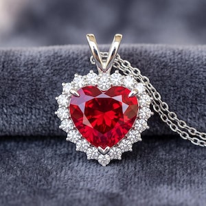 May include: A heart-shaped red gemstone pendant set in a silver-toned metal frame, surrounded by a halo of small clear stones. The pendant is attached to a delicate silver-toned chain, featuring a classic prong setting that secures the centre stone.