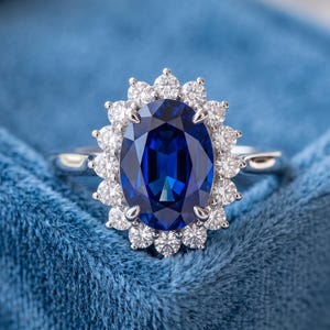 Könnte beinhalten: Ein silberfarbener Ring mit einem großen, oval geschliffenen, tiefblauen Saphir als Mittelstein, umgeben von einem Kranz aus vierzehn kleinen, runden, klaren weißen Diamanten. Der Ring hat eine polierte Metallschiene auf blauem Samt.