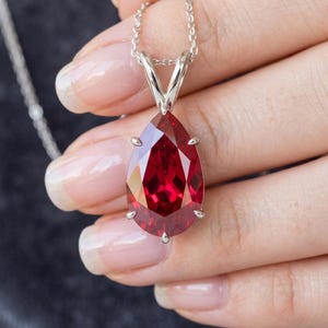 Pode incluir: Um colar de prata com um pingente de pedra preciosa vermelha facetada em forma de gota. A pedra preciosa é presa por quatro garras. A corrente é delicada e prateada. O pingente está em uma mão.