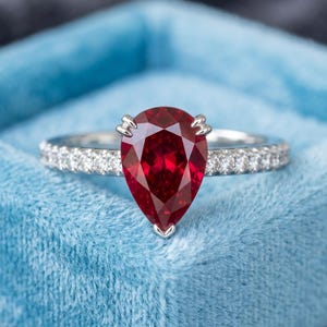 May include: A silver-coloured ring featuring a large, pear-shaped, deep red gemstone set in a prong setting. The band is slender and encrusted with a row of small, clear, round-cut stones that extend along both sides of the central red gemstone.