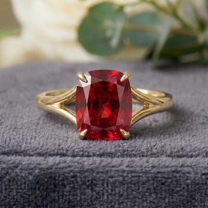 May include: A gold ring with a large, rectangular, deep red gemstone. The ring's band has a split design, and the gemstone is set with four prongs. The ring is displayed on a grey velvet surface.