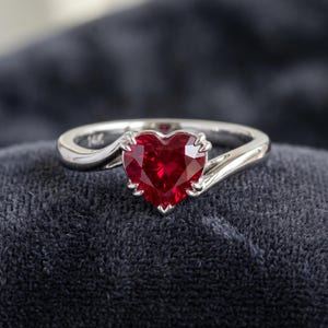 May include: A silver ring with a heart-shaped, deep red gemstone. The gemstone is the focal point, set in a prong setting. The band curves gracefully, creating a classic and elegant design. The ring is displayed on a dark gray surface.